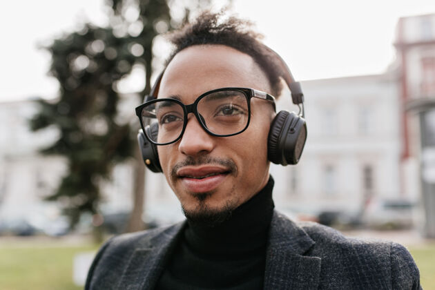 混血儿戴着眼镜的非洲男人在城市里摆出温柔的微笑戴着大耳机的漂亮黑发黑人男人在户外发冷享受帅气表情