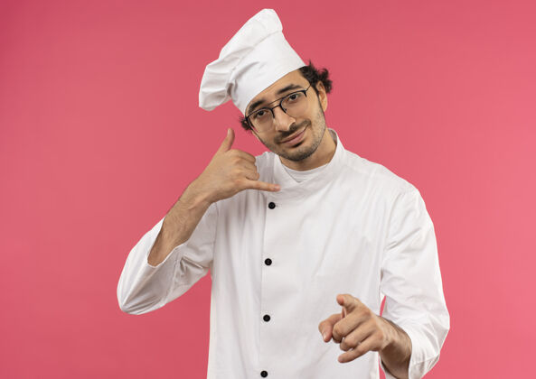 请高兴的年轻男厨师穿着厨师制服和眼镜显示来电手势与你的手势粉红色烹饪背景电话
