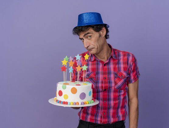 感觉可疑的中年聚会男子戴着聚会帽拿着生日蛋糕看着前面孤立的紫色墙壁生日姿势站着