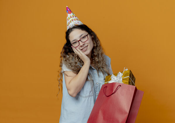 礼物微笑的年轻女孩戴着眼镜 戴着生日帽 手里拿着礼盒和袋子年轻女孩微笑