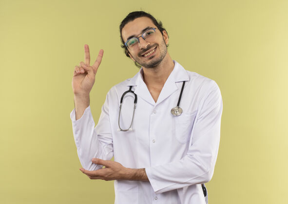 复制微笑的年轻男医生戴着眼镜 身穿白色长袍 手持听诊器 在绿色上显示和平姿态医生年轻背景