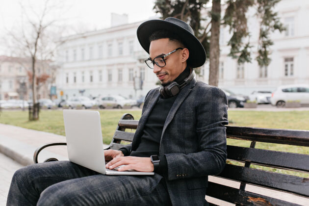 混血儿戴着帽子的自由职业者正坐在公园里 手里拿着电脑英俊的非洲年轻人在大自然的键盘上打字的户外照片设备自由职业者公园