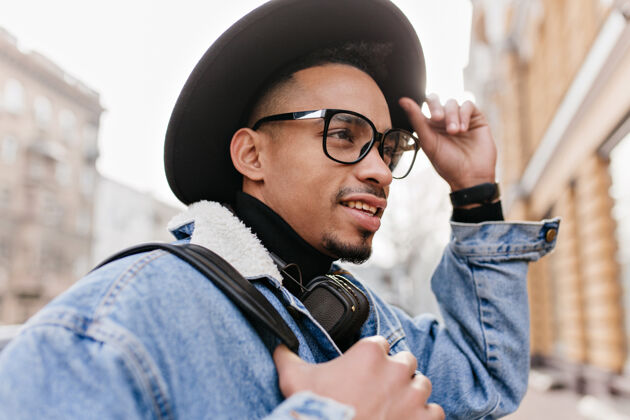 年轻充满灵感的混血儿男人穿着休闲牛仔夹克走在街上一张非洲男人戴着黑帽子和时髦眼镜在城市里度过时光的户外照片男人夹克情感