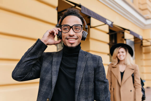 年轻穿着米色服装的优雅金发女孩站在笑眯眯的非洲男人后面戴着耳机的黑人男人在街上放松的户外照片城镇城市年轻