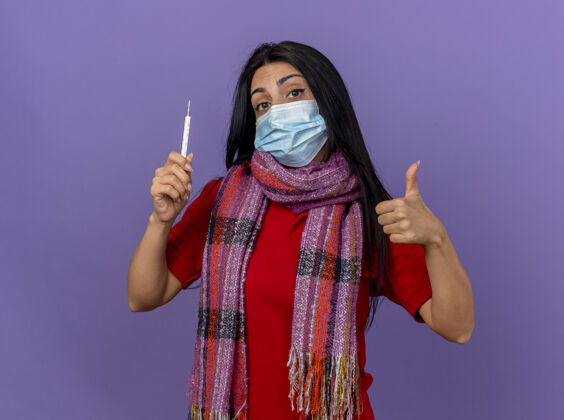 表演自信的年轻生病的女人戴着面具和围巾拿着温度计看着前面伸出大拇指孤立地站在紫色的墙上站着人脸