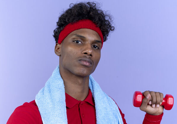 运动自信的年轻美国黑人运动男子戴着头带和手环举着哑铃 肩上挂着毛巾 隔离在蓝色的墙上毛巾哑铃腕带