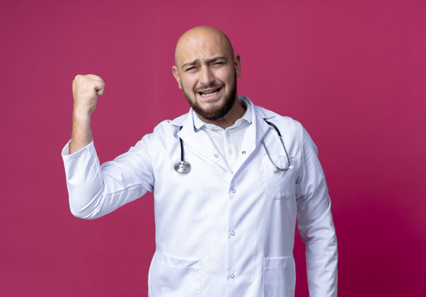 医生快乐的年轻秃头男医生穿着医用长袍和听诊器显示出强烈的手势隔离在粉红色的墙上表演长袍欢乐