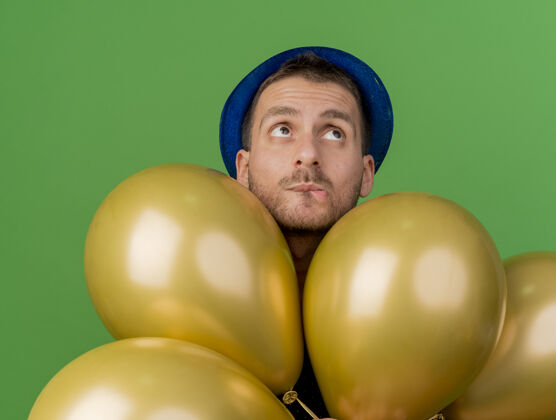 党戴着蓝色派对帽的焦急帅哥手里拿着氦气球 孤零零地看着绿色的墙上英俊姿势黄金