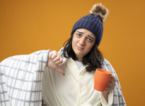 公民年轻的生病的女人穿着长袍 戴着冬天用格子布包起来的帽子 手里拿着一杯茶 看着前面 大拇指朝下 孤零零地躺在橘色的墙上抱表演茶