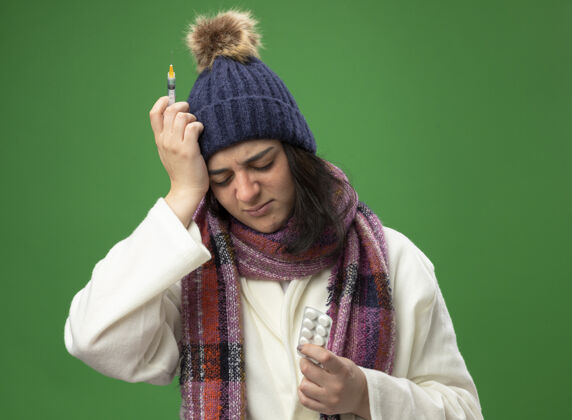 疼痛带着长袍 冬帽 围巾 拿着注射器和药片的年轻生病的女人 手放在头上 闭着眼睛 隔离在绿色的墙上帽子表情注射器
