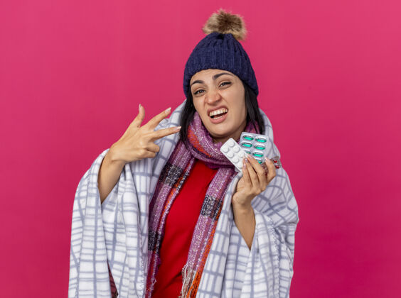 脸自信的年轻生病的女人戴着冬天的帽子 围着方格布围巾 手里拿着一包药片 看着前面 三只手被隔离在粉红色的墙上三包装格子