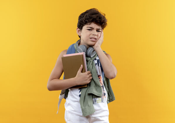 悲伤可怜的小男孩戴着书包和耳机 手里拿着书 手放在脸上 脸上泛着黄色背脸颊表情
