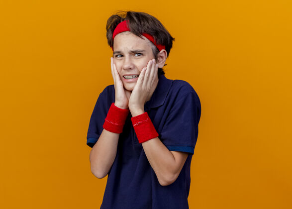 背带年轻帅气的运动男孩戴着头带和护腕 戴着牙套看着前面 手放在脸上 隔离在橙色的墙上穿着男孩年轻