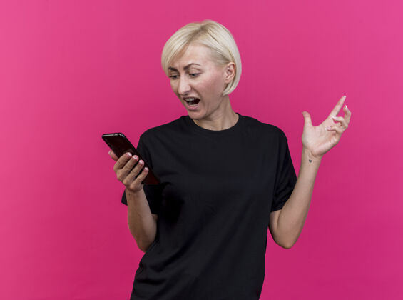 金发愤怒的中年金发女子手持手机 看着手机 手在空气中隔离在粉红色的墙上表情人空气