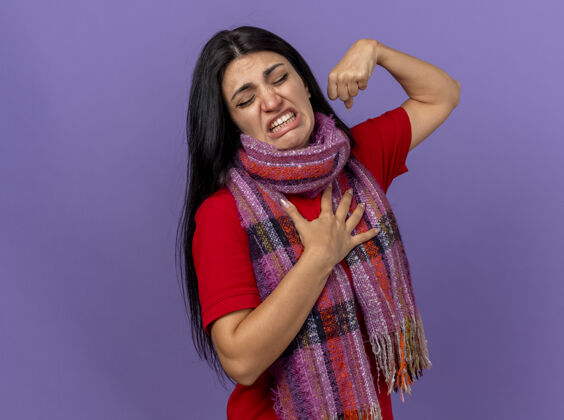 站立一个戴着围巾的疼痛的年轻女人做着一个有力的手势 把手放在胸前 闭着眼睛孤立在紫色的墙上强壮手女人