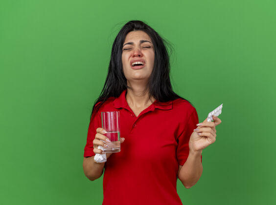 人一个年轻的生病的女人 抱着一包药片 一杯水和一张餐巾 闭着眼睛 孤零零地站在绿色的墙上拿着表情市民