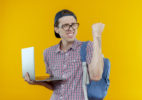 关闭闭上眼睛 快乐的年轻学生男孩戴着背包 戴着眼镜 戴着帽子 拿着笔记本电脑 在白色屏幕上显示“是”的手势背景笔记本电脑是的