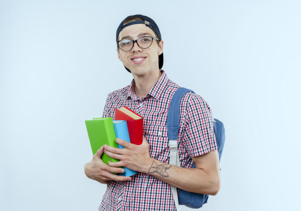 背景带着微笑的年轻学生男孩背着书包 戴着眼镜和帽子 手里拿着白色的书微笑年轻眼镜