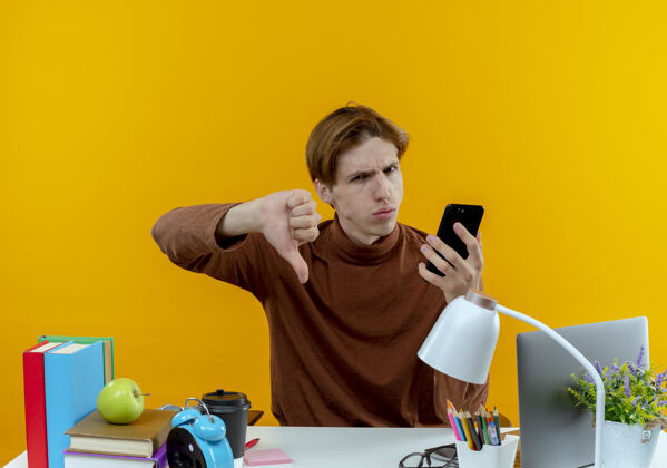 坐着坐在书桌旁拿着学习工具拿着电话的一个没出息的年轻学生 他的大拇指放在黄色的桌子上男孩按住书桌