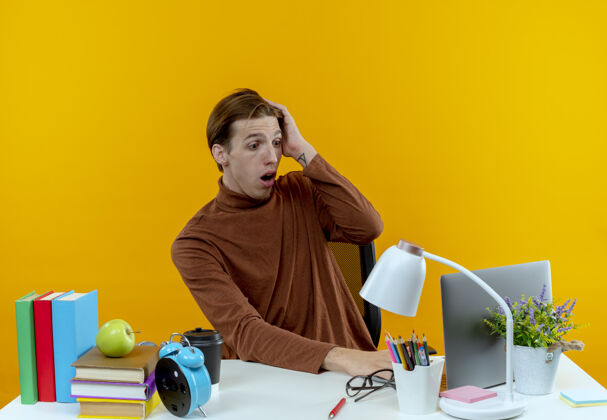 黄色惊讶的年轻学生男孩坐在书桌旁 手里拿着用过的笔记本电脑 手放在黄色的头上使用学生工具
