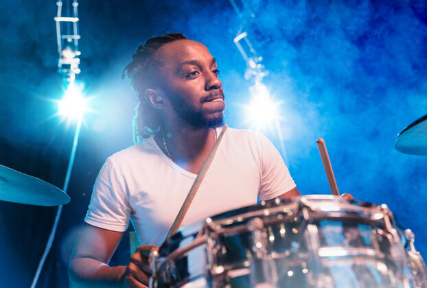 男人年轻的非洲裔美国爵士乐音乐家或鼓手 在他周围的炽热烟雾中在蓝色背景上打鼓表演鼓手时尚