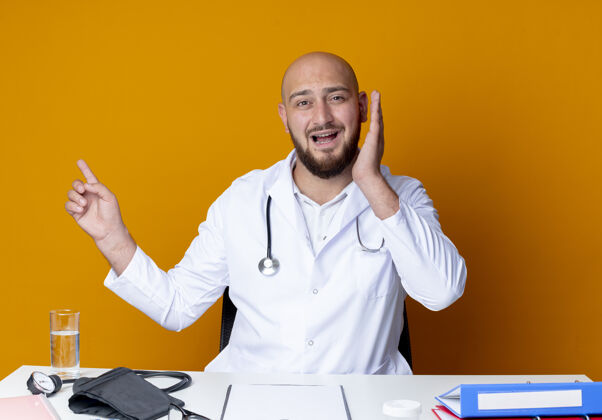 医生年轻的秃头男医生穿着医用长袍和听诊器坐在办公桌旁 吓得要命橙色听诊器桌子
