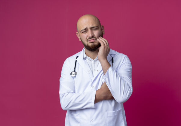 粉色悲伤的年轻男医生穿着医用长袍和听诊器把手放在疼痛的牙齿上隔离在粉红色男性医生长袍