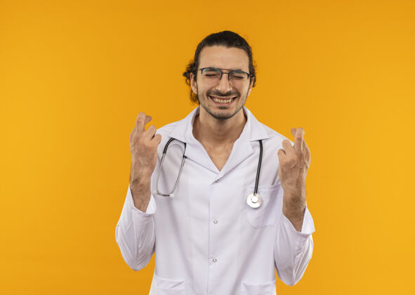 请年轻的医生戴着医用眼镜 穿着医用长袍 戴着听诊器 双手交叉着手指 眼睛闭着 很高兴黄色医学眼睛