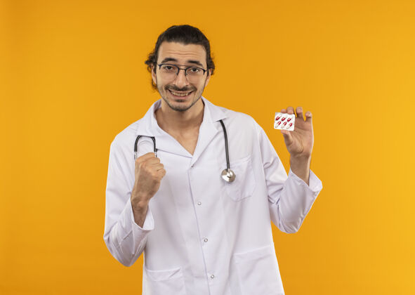 手势微笑着的年轻医生戴着医用眼镜穿着医用长袍手持听诊器拿着药片 在黄色上显示“是”的手势背景听诊器黄色
