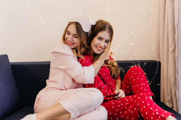 沙发一个穿着粉色睡衣的女孩坐在蓝色沙发上欣喜若狂一个穿着红色睡衣的黑发女人在笑 而她的姐姐在拥抱她室内娱乐周末