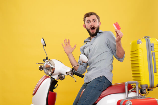 车辆旅游概念的正面视图 一个情绪激动的年轻人坐在摩托车上 提着手提箱 手里拿着黄色的银行卡自行车手提箱摩托车
