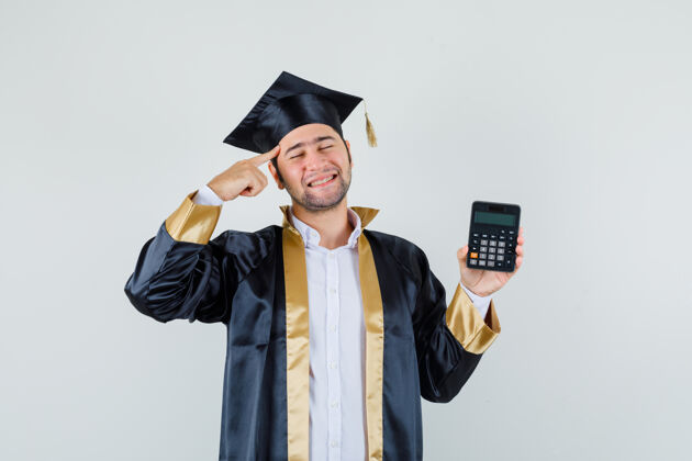 自信身穿毕业制服的年轻人拿着计算器 手指放在太阳穴上 看上去很高兴 正对着前方看计算器毕业站