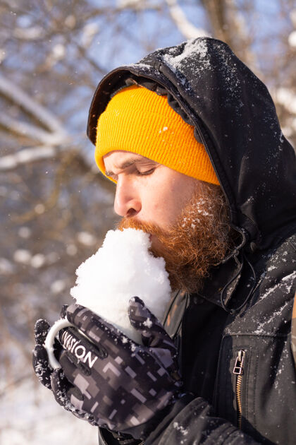 人在一个阳光明媚的日子里 一个戴着橙色明亮帽子的残暴的成年男子拿着装满雪的杯子在雪林里玩自然毛皮脸
