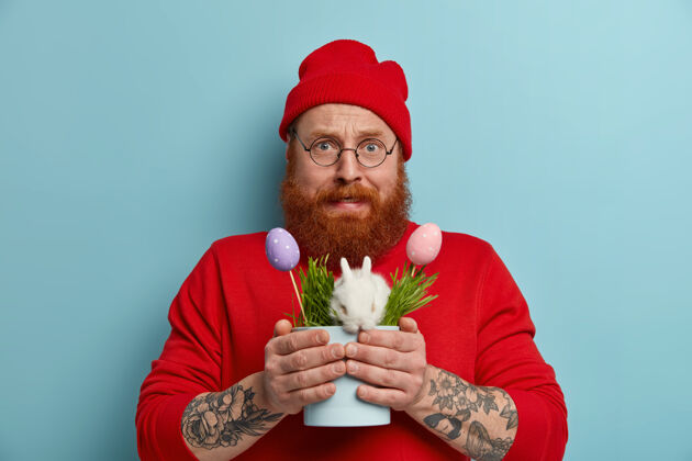 草紧张的大胡子男人准备复活节庆典 手里拿着小白兔和彩蛋的罐子 一脸困惑 穿着红色的衣服 在室内摆姿势春节假期兔子紧张传统