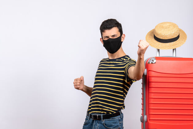 人正面图幸福的年轻游客戴着黑色面具站在红色手提箱旁展示他的拳头站手提箱帅哥