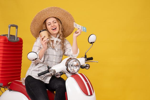 摩托车微笑的年轻女子戴着帽子 坐在摩托车上 手里拿着咖啡和黄色的车票高尔夫车辆年轻女子