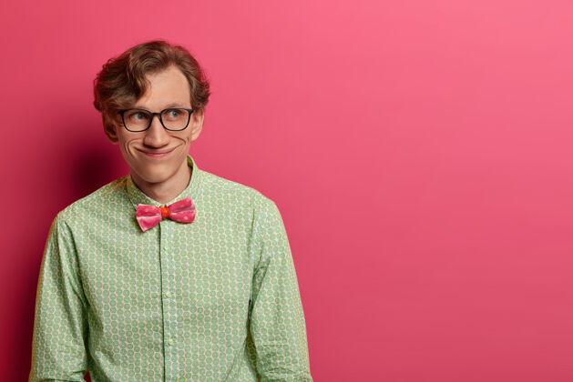 微笑有趣的快乐男人的照片穿着优雅的绿色衬衫和蝴蝶结 戴着透明的眼镜 有着愉快的积极的目光 计划着一些事情 孤立在粉红色的墙上 文本的复制空间老师男性人