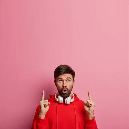 面部迷人印象深刻的胡须男子梅洛曼点以上 显示一些惊人的向上 使用立体声耳机听音乐 喘息的奇迹 隔离在粉红色的墙壁促销概念不刮胡子惊呆男性