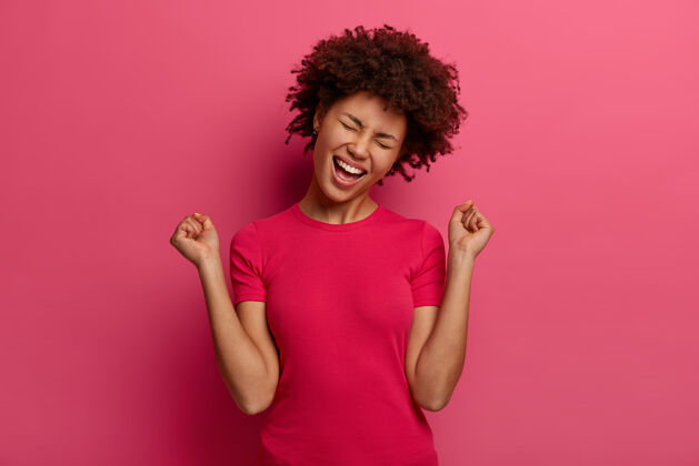 握紧人 情感 胜利和成功的概念快乐的美国黑人少女庆祝胜利 举起拳头 有乐观的情绪 穿着休闲服 孤立在粉红色的墙上高兴幸运美国