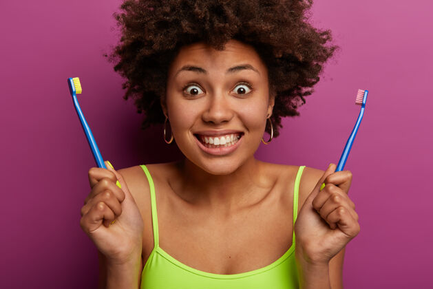 种族卷发女性短镜头表情愉快 手持两把牙刷 露出洁白完美的牙齿 有每天卫生的晨间程序 健康的皮肤 隔离在紫色的墙壁上微笑口腔肖像