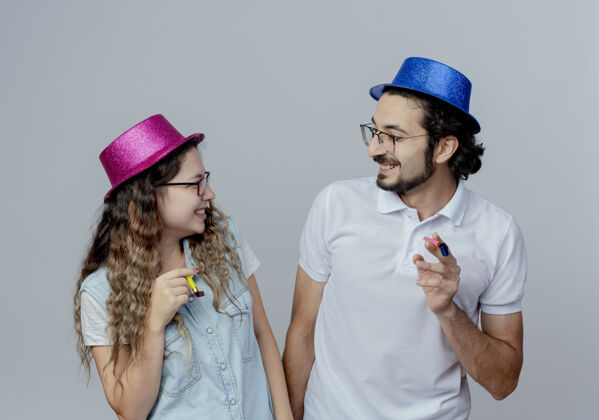 戴着微笑着的年轻夫妇戴着粉色和蓝色的帽子看着对方 手持哨子孤立地站在白色的舞台上抱着年轻情侣