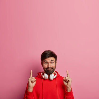 手指留着胡子的时髦男人把食指指向上方 显示出惊人的空白 给出很好的报价 紧闭嘴唇 戴着立体声耳机和红色连帽衫 隔离在粉色粉彩墙上向上看向上个人模型