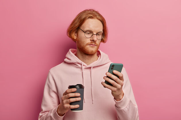 男性严肃的大胡子男人使用现代手机进行在线交流 查看电子邮件 专注于屏幕 喝外卖咖啡 戴眼镜和连帽衫 隔离在粉红色的墙上严肃男人电话