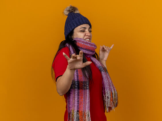 衣服愤怒的年轻生病的女人戴着冬天的帽子和围巾站在侧视图保持双手在空气中闭着眼睛隔离在橙色的墙上恼怒冬天空气