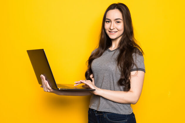 室内一个微笑的少女拿着笔记本电脑隔离在黄色墙上的肖像欢呼女性微笑