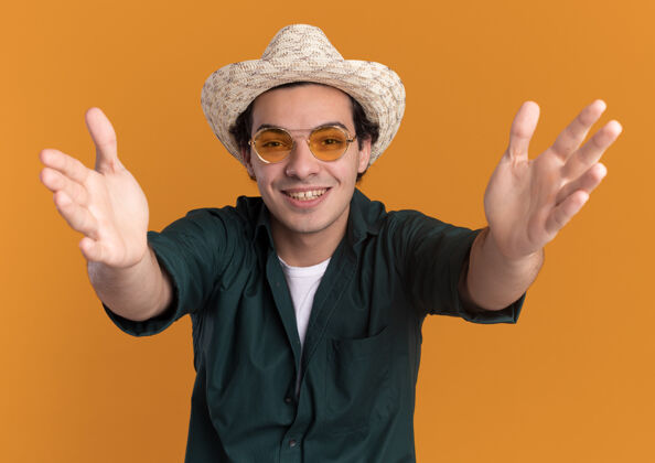 快乐身穿绿色衬衫 戴着夏季帽子 戴着眼镜 脸上带着微笑站在橙色的墙上的快乐的年轻人眼镜夏天衬衫
