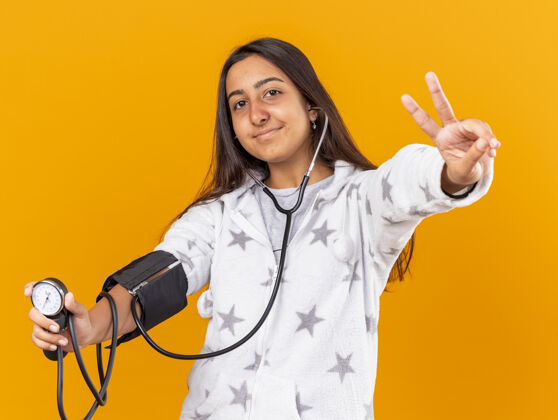 女孩高兴的年轻病女孩测量自己的压力血压计显示和平姿态隔离在黄色背景上压力和平疾病