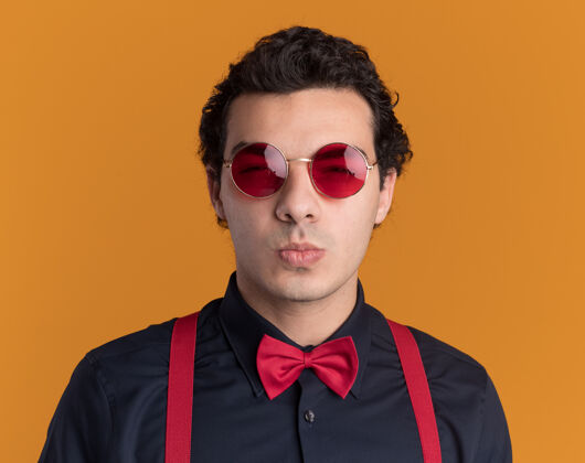 表情带着蝴蝶结戴着眼镜和吊带的时髦男人站在橙色的墙上 带着怀疑的表情看着前面时尚立场眼镜