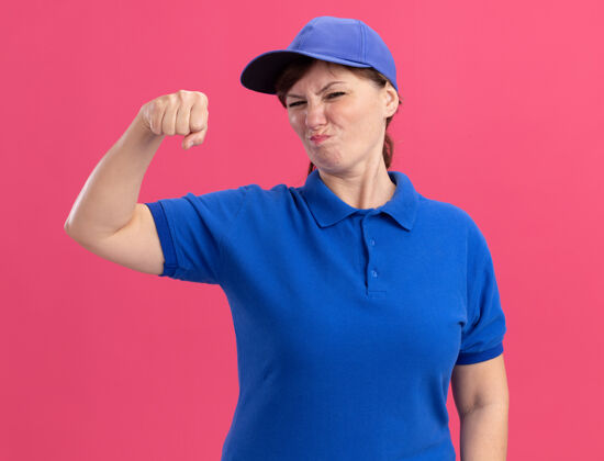 显示身穿蓝色制服 头戴鸭舌帽的中年女送货员站在粉红色的墙上 看着前面紧握的拳头 不高兴地站在那里立场中间帽子