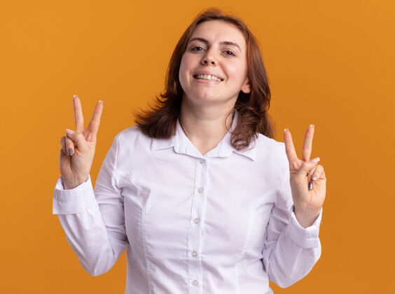 女人身穿白衬衫的年轻女子看着前面 开心而积极地微笑着 橙色的墙上挂着v字标志Vsign快乐展示